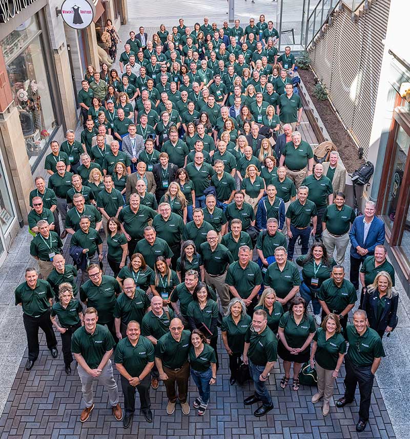 A large group of people in green shirts stands in a courtyard, forming a V-shape, reminiscent of a team of business brokers strategizing their next big deal, all facing the camera.